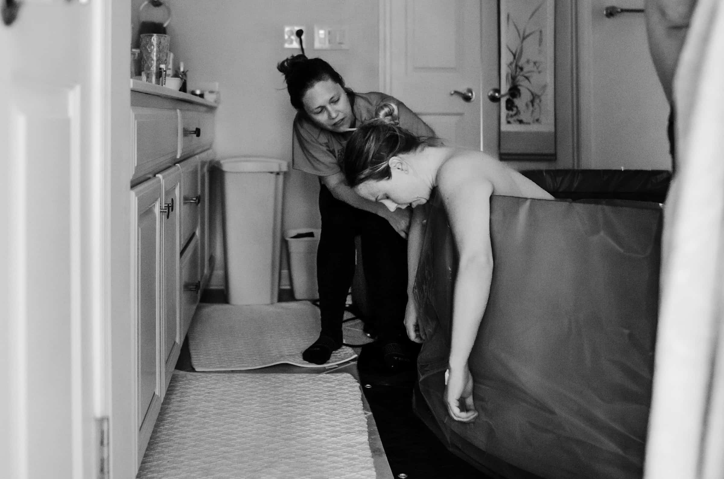 A pregnant woman supported by a companion in a softly lit bathroom